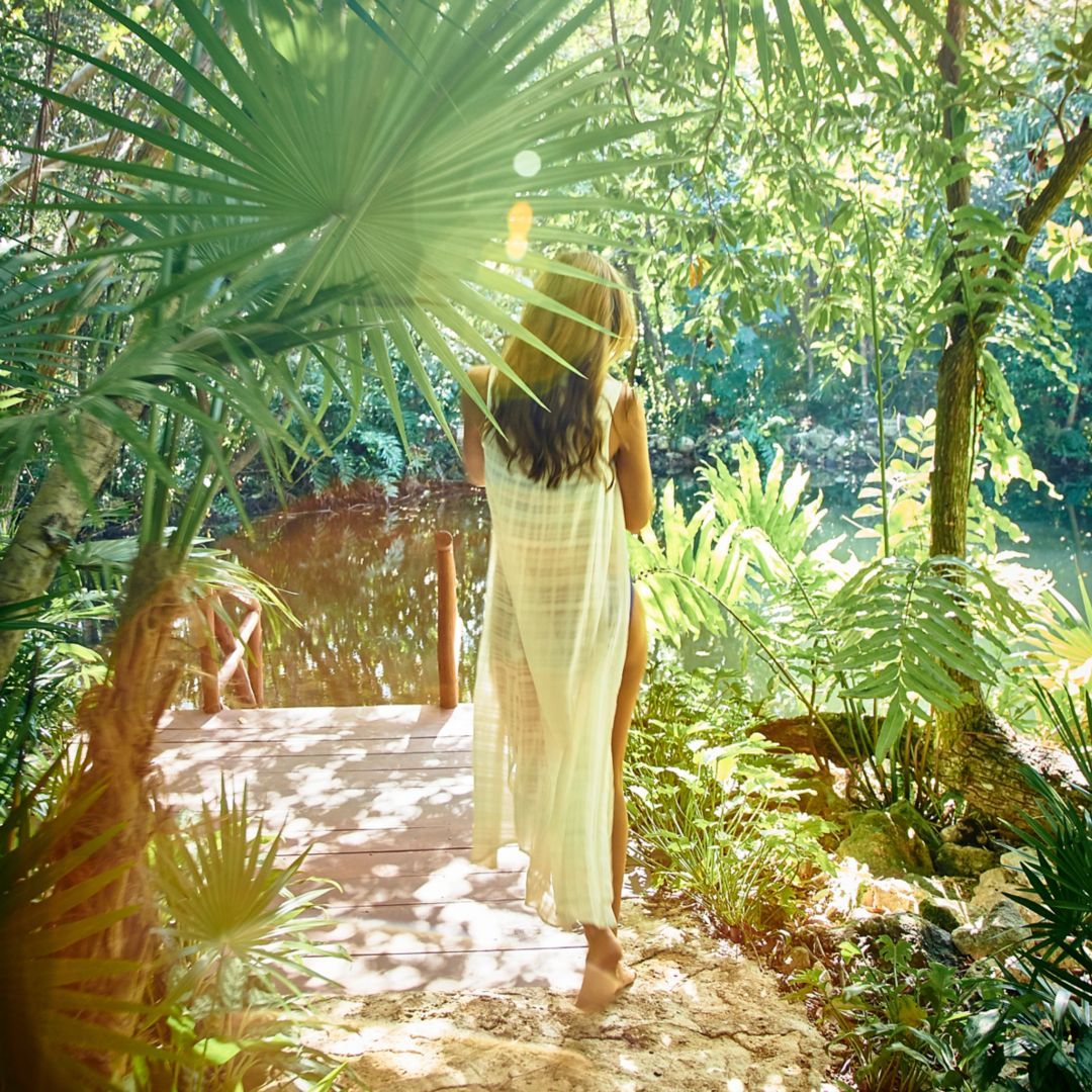 Woman with brown hair and white coverup walks barefoot through jungle on stone path in afternoon sunlight